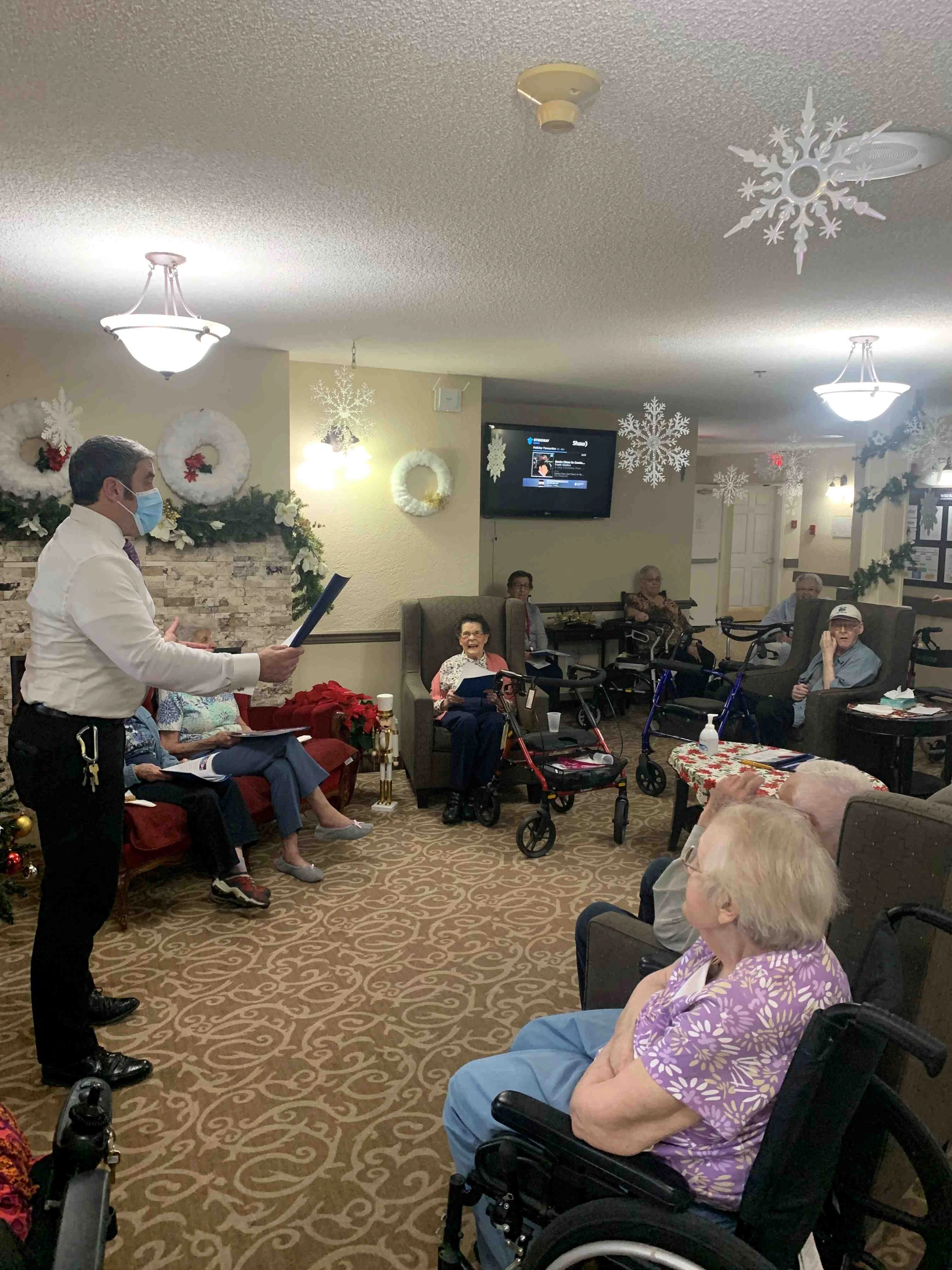 A group of elderly people sitting in a senior living common room on Christmas Eve and a man speaking